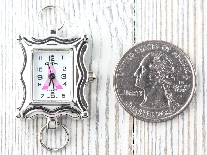 K&ouml;nnte beinhalten: Eine silberfarbene Geneva-Quarzuhr mit rosa Schleifendesign auf dem Zifferblatt, neben einer M&uuml;nze. Die Uhr hat ein rechteckiges Zifferblatt mit schwarzen Ziffern und Zeigern. Die Uhr ist etwa 2.5 cm lang.