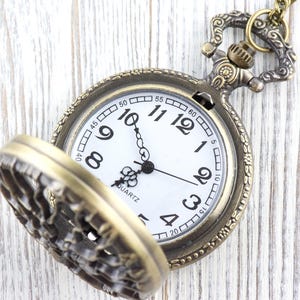 May include: A vintage pocket watch in a bronze tone, with a white clock face and black numerals. The watch hands point to ten past ten. The watch lid is open, revealing the detailed design. The word "QUARTZ" is printed on the face.