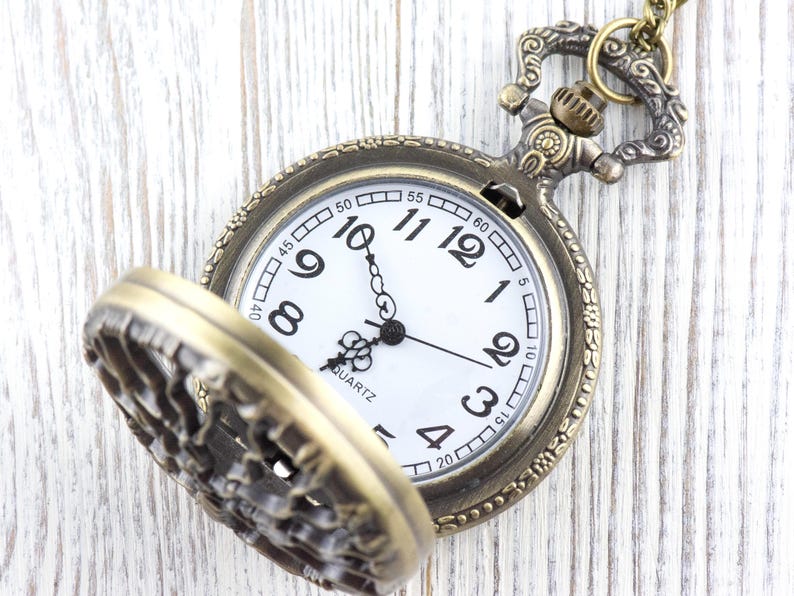 May include: A vintage pocket watch in a bronze tone, with a white clock face and black numerals. The watch hands point to ten past ten. The watch lid is open, revealing the detailed design. The word "QUARTZ" is printed on the face.