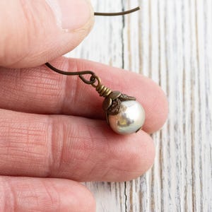May include: A single pearl earring with a bronze-colored hook and decorative cap. The pearl is a creamy white color and has a slight luster. The earring is held in a person's hand against a white wooden background.