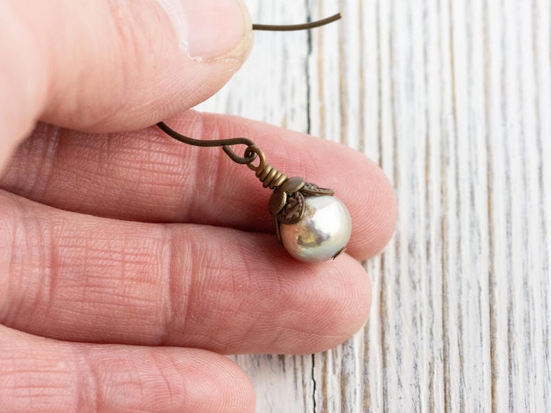 May include: A single pearl earring with a bronze-colored hook and decorative cap. The pearl is a creamy white color and has a slight luster. The earring is held in a person's hand against a white wooden background.