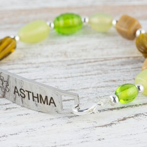 May include: A silver medical alert bracelet with the word "ASTHMA" engraved on it. The bracelet is made with green, brown, and beige beads.