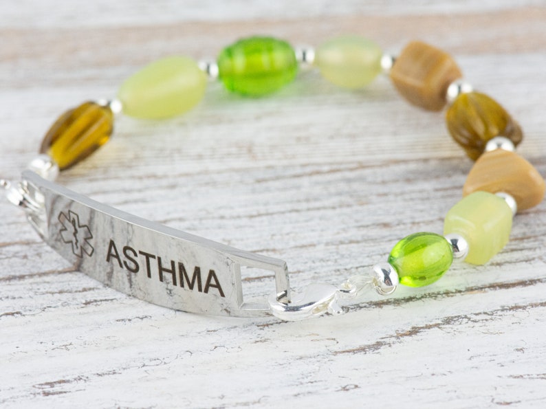 May include: A silver medical alert bracelet with the word "ASTHMA" engraved on it. The bracelet is made with green, brown, and beige beads.