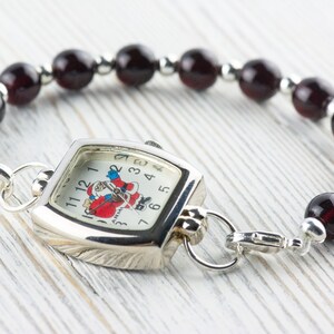 May include: A silver watch with a square face and a Santa Claus design. The watch is attached to a bracelet made of dark red beads.