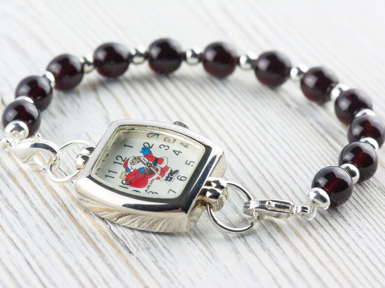 May include: A silver watch with a square face and a Santa Claus design. The watch is attached to a bracelet made of dark red beads.