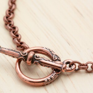 Puede incluir: Un collar de cadena de cobre con un cierre de palanca decorativo. El cierre es ornamentado y presenta un dise&ntilde;o detallado.