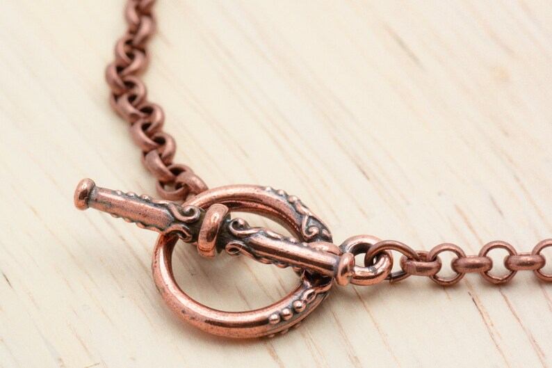 Puede incluir: Un collar de cadena de cobre con un cierre de palanca decorativo. El cierre es ornamentado y presenta un dise&ntilde;o detallado.