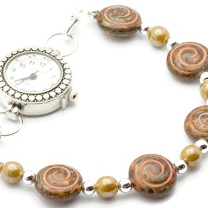 May include: A silver watch bracelet with a white watch face and a decorative bezel. The bracelet features brown and orange swirl beads, olive green beads, and silver spacer beads. The watch face displays the time.
