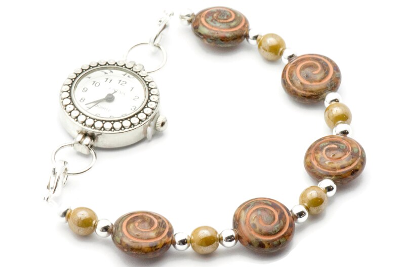 May include: A silver watch bracelet with a white watch face and a decorative bezel. The bracelet features brown and orange swirl beads, olive green beads, and silver spacer beads. The watch face displays the time.