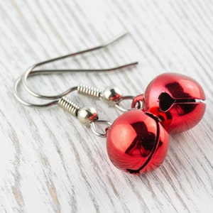May include: A pair of festive red bell earrings. Each earring features a shiny, round bell with a slit and a silver-colored hook. The earrings are displayed on a white, weathered wooden surface.