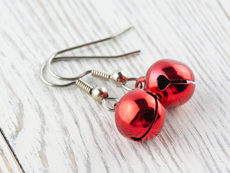 May include: A pair of festive red bell earrings. Each earring features a shiny, round bell with a slit and a silver-colored hook. The earrings are displayed on a white, weathered wooden surface.