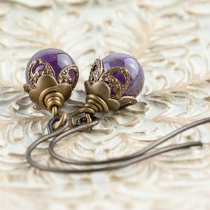 K&ouml;nnte beinhalten: Ein Paar Ohrh&auml;nger mit tiefvioletten, kugelf&ouml;rmigen Perlen. Die Perlen sind in aufwendigen, antik bronzefarbenen Metallarbeiten eingefasst. Die Ohrringe haben ein florales Design an der Oberseite und einen einfachen Hakenverschluss.