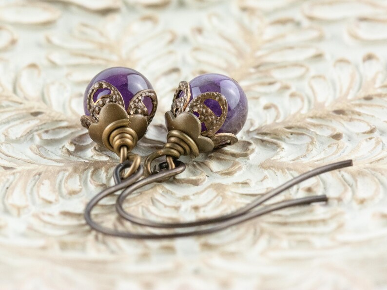 K&ouml;nnte beinhalten: Ein Paar Ohrh&auml;nger mit tiefvioletten, kugelf&ouml;rmigen Perlen. Die Perlen sind in aufwendigen, antik bronzefarbenen Metallarbeiten eingefasst. Die Ohrringe haben ein florales Design an der Oberseite und einen einfachen Hakenverschluss.
