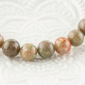 Pu&ograve; includere: Primo piano di un braccialetto con perline rotonde in pietra naturale. Le perline presentano un mix di colori, tra cui verde, marrone e rosso, con un motivo maculato. Il braccialetto &egrave; probabilmente realizzato con elastico o materiale simile.