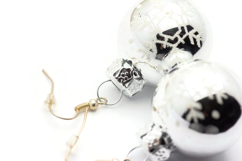 May include: A pair of silver Christmas ornament earrings with snowflake designs. The earrings are attached to gold-toned hooks.