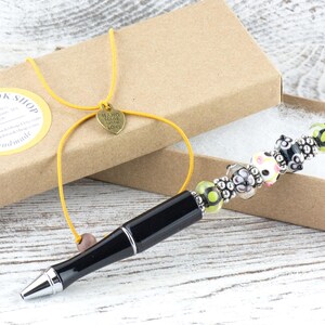 Op de afbeelding: Een zwarte pen met een decoratief kralengedeelte, gepresenteerd in een bruine doos. De kralen zijn in verschillende kleuren, waaronder roze, groen en zwart. De doos is vastgebonden met een geel koord en een hartvormige bedel.