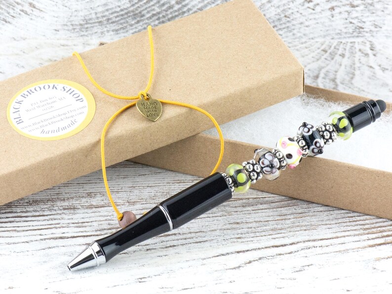 Op de afbeelding: Een zwarte pen met een decoratief kralengedeelte, gepresenteerd in een bruine doos. De kralen zijn in verschillende kleuren, waaronder roze, groen en zwart. De doos is vastgebonden met een geel koord en een hartvormige bedel.