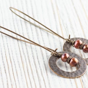 May include: A pair of bronze-colored earrings with a hammered oval design and three reddish-brown beads. The earrings have long, thin ear wires. The jewelry is displayed on a white wooden surface.