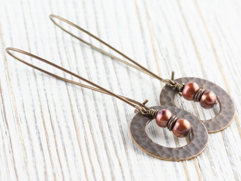 May include: A pair of bronze-colored earrings with a hammered oval design and three reddish-brown beads. The earrings have long, thin ear wires. The jewelry is displayed on a white wooden surface.