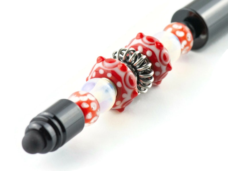 Puede incluir: Un bol&iacute;grafo de punta de aguja negro con una cuenta de metal plateado y cuentas de vidrio rojo y blanco.