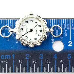 May include: A silver-toned quartz watch face with a white dial and black numerals. The watch face is surrounded by a decorative bezel with clear crystals. The watch face is approximately 1 inch in diameter. The watch is on a blue ruler.