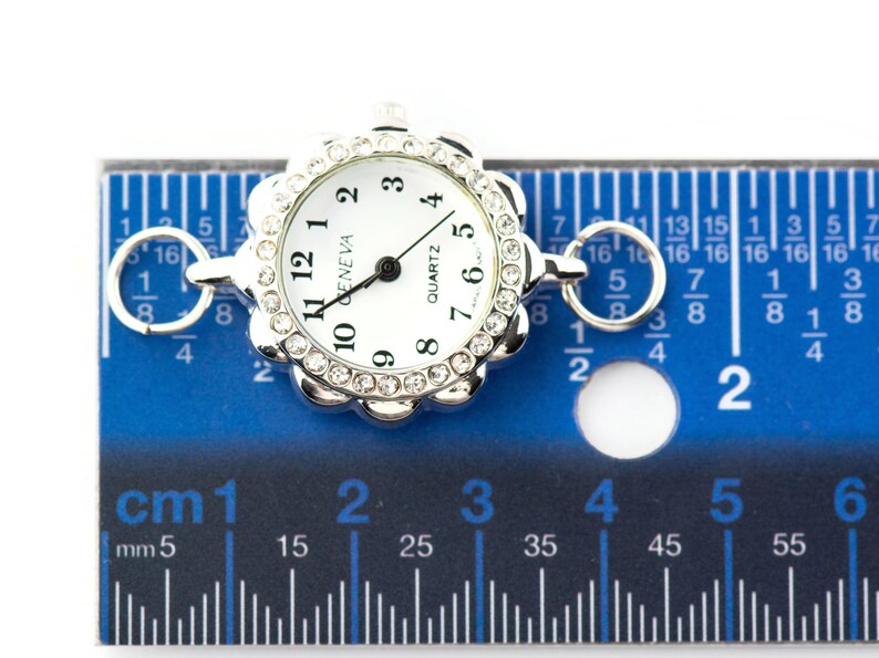 May include: A silver-toned quartz watch face with a white dial and black numerals. The watch face is surrounded by a decorative bezel with clear crystals. The watch face is approximately 1 inch in diameter. The watch is on a blue ruler.