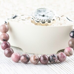 May include: A pink and black gemstone bracelet with a silver watch attached. The bracelet is made of round beads and the watch has a white face with silver hands and a crystal-encrusted bezel.