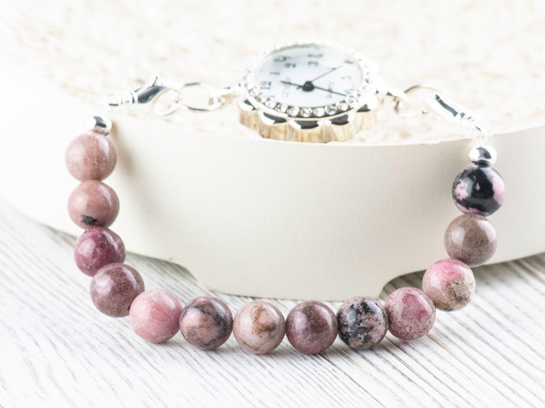 May include: A pink and black gemstone bracelet with a silver watch attached. The bracelet is made of round beads and the watch has a white face with silver hands and a crystal-encrusted bezel.