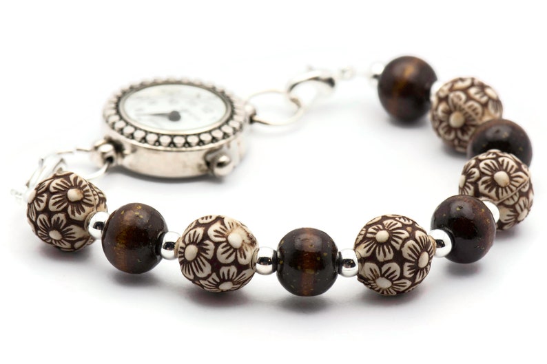 Op de afbeelding: Een zilveren horloge met een witte wijzerplaat en zwarte wijzers bevestigd aan een bruin kralenarmband. De kralen hebben een bloemmotief.