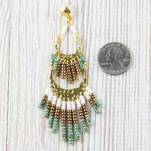 May include: Gold tone dangle earrings with white, brown, and turquoise beads. The earrings are about the size of a US quarter.