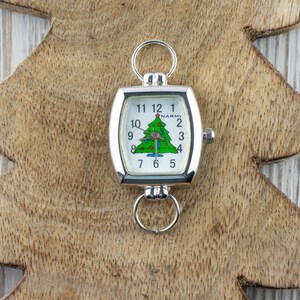May include: A silver-toned watch with a white face featuring a green Christmas tree design. The watch has a rectangular shape with rounded edges and a ring at the top and bottom. The watch is set against a wooden Christmas tree-shaped background.