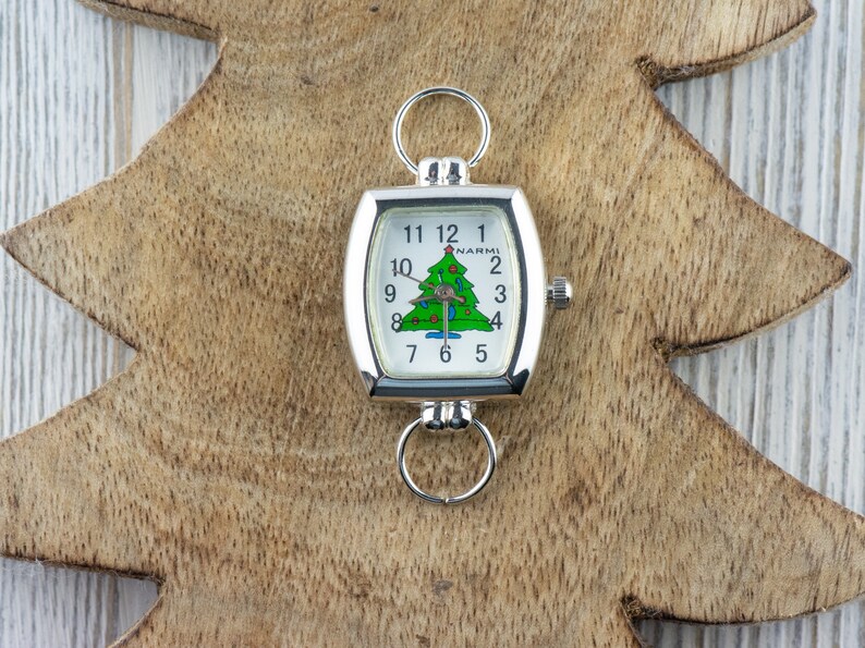 May include: A silver-toned watch with a white face featuring a green Christmas tree design. The watch has a rectangular shape with rounded edges and a ring at the top and bottom. The watch is set against a wooden Christmas tree-shaped background.