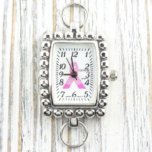 May include: Silver-toned rectangular watch with a white face featuring black numerals and hands. A pink ribbon graphic is centered on the face. The watch is surrounded by a beaded border and has a ring at the top and bottom.