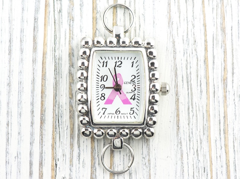 May include: Silver-toned rectangular watch with a white face featuring black numerals and hands. A pink ribbon graphic is centered on the face. The watch is surrounded by a beaded border and has a ring at the top and bottom.