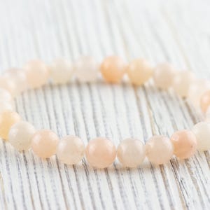 Op de afbeelding: Een armband gemaakt van lichtroze en witte ronde kralen. De armband ligt op een wit houten oppervlak.
