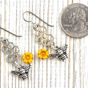 May include: Silver-tone bee earrings with honeycomb and yellow flower accents. The earrings feature detailed bee charms and are displayed next to a coin for size reference. The earrings are on a white wooden surface.