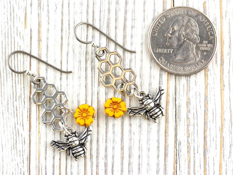 May include: Silver-tone bee earrings with honeycomb and yellow flower accents. The earrings feature detailed bee charms and are displayed next to a coin for size reference. The earrings are on a white wooden surface.