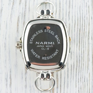 Pu&ograve; includere: Un orologio argentato con quadrante rettangolare. Il retro dell'orologio &egrave; nero con testo bianco che recita "STAINLESS STEEL BACK", "NARMI JAPAN MOVT CL-9" e "WATER RESISTANT". L'orologio ha un anello in alto e in basso.