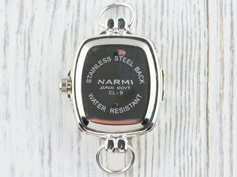 Pu&ograve; includere: Un orologio argentato con quadrante rettangolare. Il retro dell'orologio &egrave; nero con testo bianco che recita "STAINLESS STEEL BACK", "NARMI JAPAN MOVT CL-9" e "WATER RESISTANT". L'orologio ha un anello in alto e in basso.