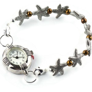 K&ouml;nnte beinhalten: Eine silberne Uhr mit einem runden Zifferblatt und r&ouml;mischen Ziffern. Die Uhr ist an einer silbernen Kette mit braunen Perlen und silbernen Seestern-Anh&auml;ngern befestigt.