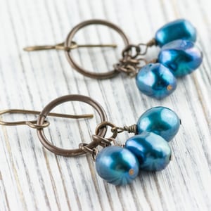 K&ouml;nnte beinhalten: Ein Paar handgefertigte Ohrringe mit einem bronzefarbenen Metalldesign. Jeder Ohrring hat ein kreisf&ouml;rmiges Element mit einem Haken und eine Ansammlung von drei irisierenden blau-gr&uuml;nen Perlen. Auf einer wei&szlig;en Holzoberfl&auml;che.