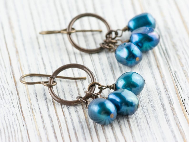 K&ouml;nnte beinhalten: Ein Paar handgefertigte Ohrringe mit einem bronzefarbenen Metalldesign. Jeder Ohrring hat ein kreisf&ouml;rmiges Element mit einem Haken und eine Ansammlung von drei irisierenden blau-gr&uuml;nen Perlen. Auf einer wei&szlig;en Holzoberfl&auml;che.