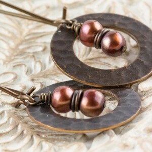 May include: A pair of bronze-toned earrings. Each earring features a hammered, circular pendant with a pair of dark pink pearls. The earrings are connected by thin, bronze-colored wires. The background is a light, textured surface.