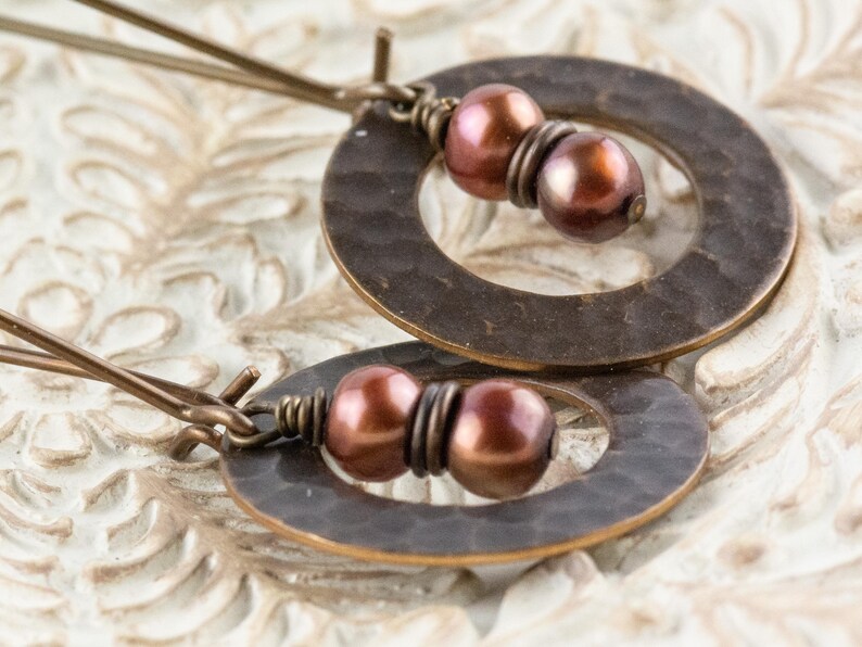 May include: A pair of bronze-toned earrings. Each earring features a hammered, circular pendant with a pair of dark pink pearls. The earrings are connected by thin, bronze-colored wires. The background is a light, textured surface.