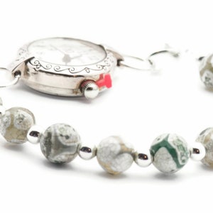 Op de afbeelding: Een zilveren horloge met een witte wijzerplaat en zwarte wijzers is bevestigd aan een armband gemaakt van grijze en groene agaat kralen.