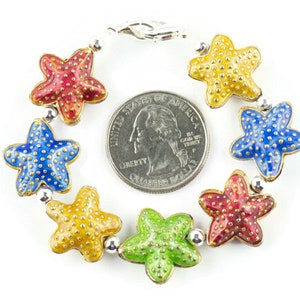 Puede incluir: Un brazalete de plata con cierre de langosta presenta siete abalorios de estrella de mar de esmalte de colores. Cada estrella de mar es de un color diferente: rojo, azul, amarillo, verde y dos estrellas de mar rojas m&aacute;s. El brazalete es aproximadamente del tama&ntilde;o de una moneda de 25 centavos de d&oacute;lar estadounidense.