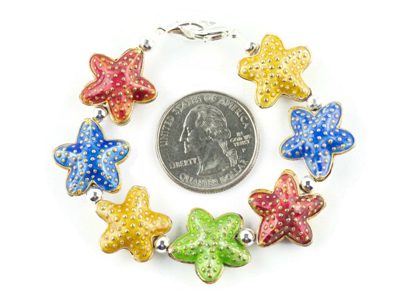 Puede incluir: Un brazalete de plata con cierre de langosta presenta siete abalorios de estrella de mar de esmalte de colores. Cada estrella de mar es de un color diferente: rojo, azul, amarillo, verde y dos estrellas de mar rojas m&aacute;s. El brazalete es aproximadamente del tama&ntilde;o de una moneda de 25 centavos de d&oacute;lar estadounidense.