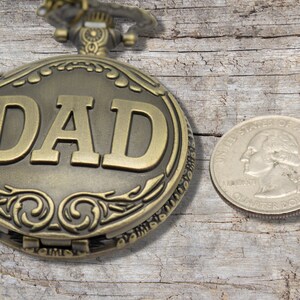 Peut inclure: Une montre de poche dor&eacute;e avec le mot "DAD" grav&eacute; sur le devant. La montre est pos&eacute;e sur une surface en bois &agrave; c&ocirc;t&eacute; d'une pi&egrave;ce de 25 cents am&eacute;ricaine.