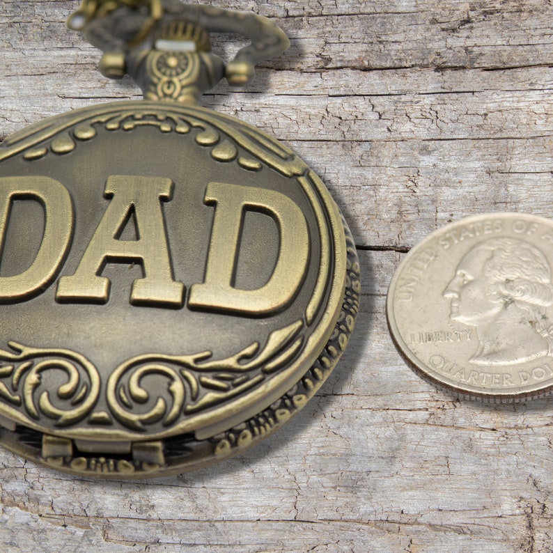 Peut inclure: Une montre de poche dor&eacute;e avec le mot "DAD" grav&eacute; sur le devant. La montre est pos&eacute;e sur une surface en bois &agrave; c&ocirc;t&eacute; d'une pi&egrave;ce de 25 cents am&eacute;ricaine.