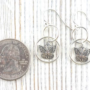 May include: Silver butterfly earrings with a quarter coin for size comparison. The earrings feature a detailed butterfly design within a circular frame, suspended from a hook. The background is a white wooden surface.